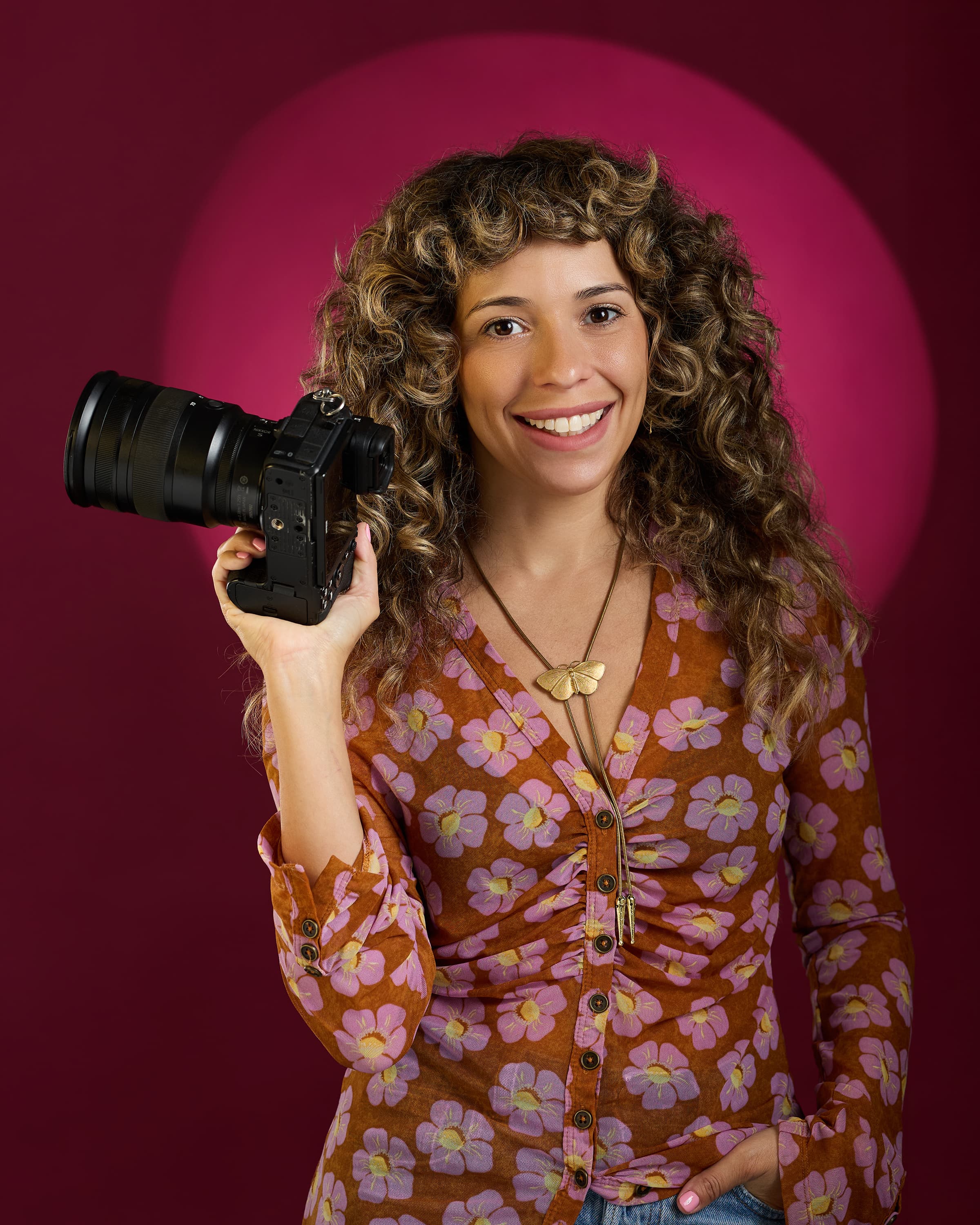 Stephanie Cousino smiling and holding her camera in front of a pink spotlight backdrop