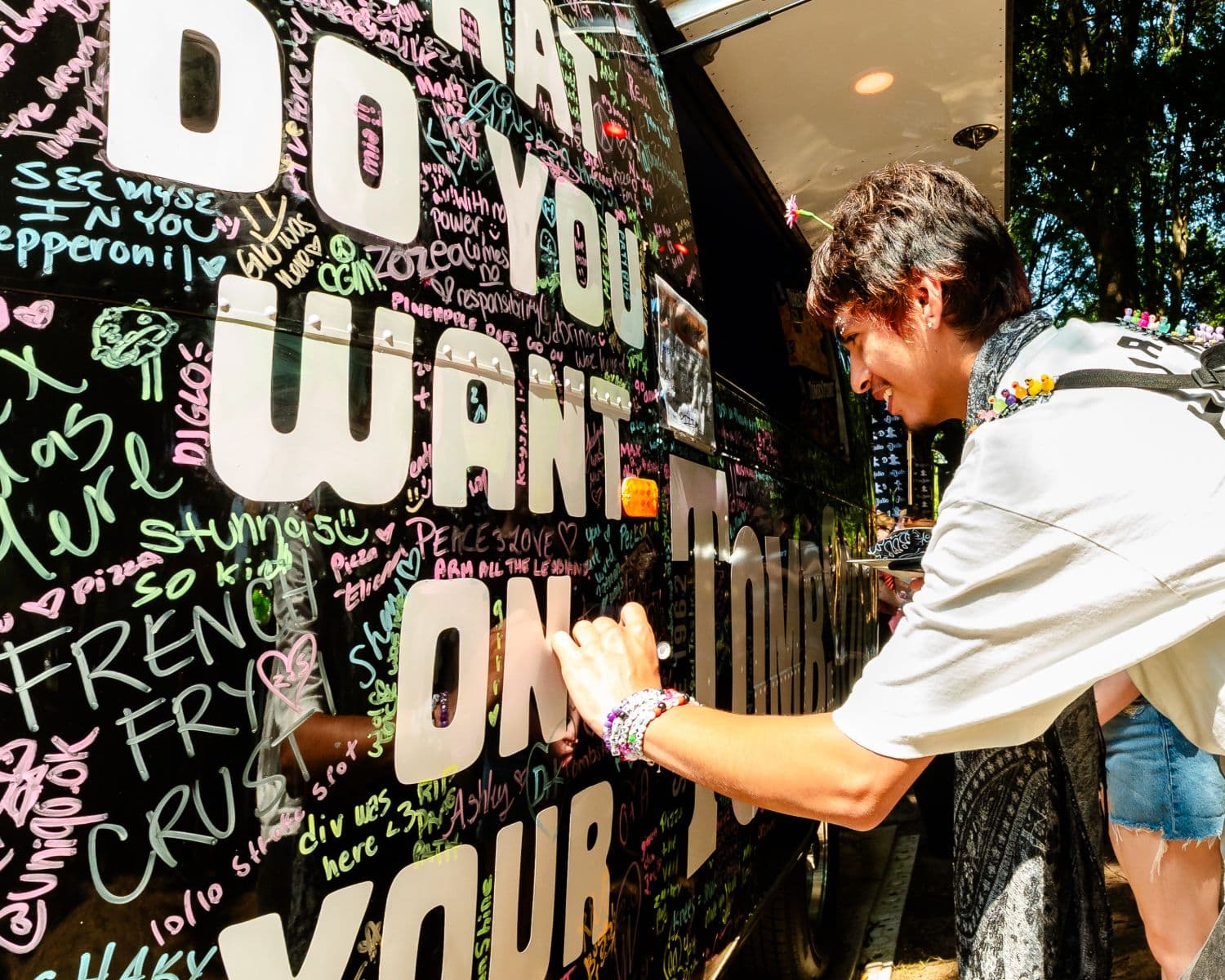 Festival goer writing with a marker on the Tombstone Pizza vehicle covered in colorful messages that reads What Do You Want On Your Tombstone