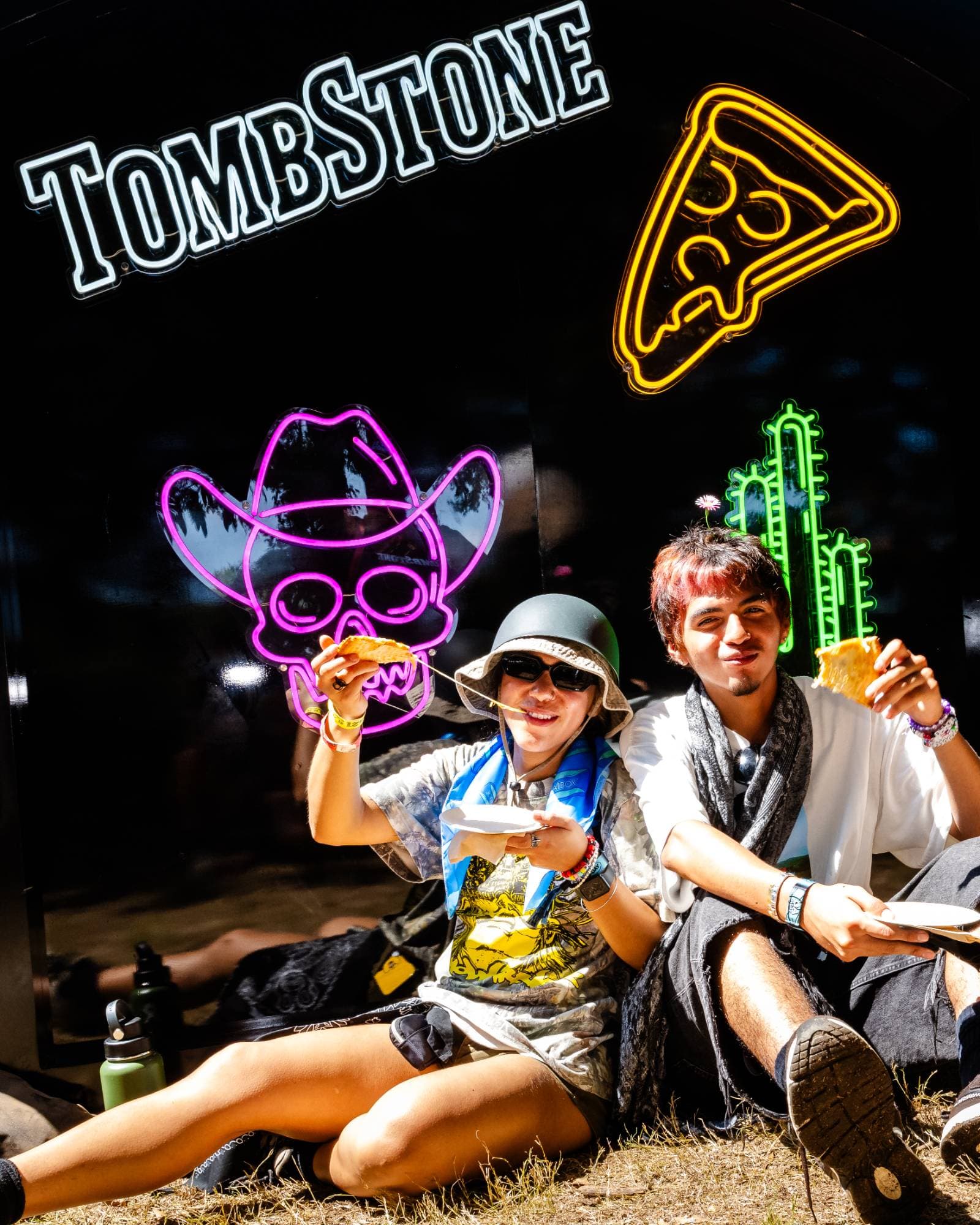 Two friends sitting in front of the Tombstone neon sign photo op with a cowboy skull, pizza slice, and cactus, holding up pizza at Shaky Knees
